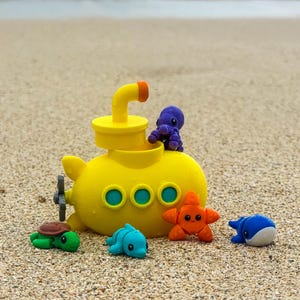 May include: A yellow submarine toy with orange accents and a propeller, surrounded by small, colorful sea creature toys, including a turtle, dolphin, starfish, octopus, and whale, all resting on a sandy beach.