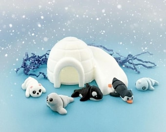 Könnte beinhalten: Eine skurrile Winterlandschaft mit einem weißen Iglu und kleinen Tierfiguren, darunter Robben, Pinguine und ein Orca, vor einem schneebedeckten blauen Hintergrund. Die Figuren sind detailliert und farbenfroh und schaffen ein verspieltes Winterthema.