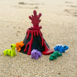 May include: A toy volcano with red lava and a black base, surrounded by colourful dinosaur figurines in green, orange, blue, purple, and green. The scene is set on a sandy beach with the ocean in the background.