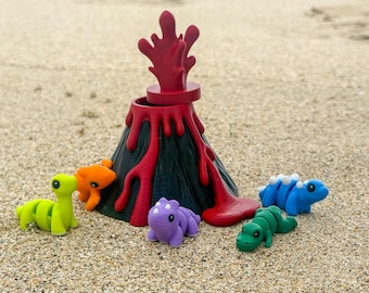 Könnte beinhalten: Ein Spielzeugvulkan mit roter Lava und schwarzer Basis, umgeben von bunten Dinosaurierfiguren in Grün, Orange, Blau, Lila und Grün. Die Szene spielt sich an einem Sandstrand mit dem Meer im Hintergrund ab.