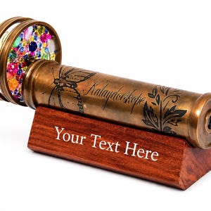 May include: A brass kaleidoscope with an etched butterfly and floral design, featuring the word "Kalaydeoscope" on the barrel. The object rests on a dark wooden stand with the engraved text "Your Text Here" and has a circular end filled with colorful beads.