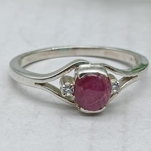 May include: A silver ring featuring a central oval-shaped deep pink ruby gemstone held in a prong setting, flanked by two small clear round accent stones on either side of the split shank band, showcasing a polished metallic finish throughout.