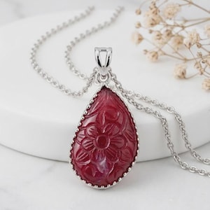 Peut inclure: Un pendentif en forme de goutte orné d'une pierre rouge foncé avec un motif floral sculpté, serti dans un métal argenté. Le pendentif est suspendu à une chaîne assortie en métal argenté, présentant une finition polie et des détails précis.