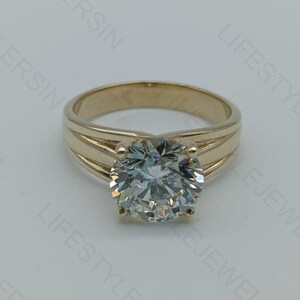 May include: A gold ring featuring a large, round, clear gemstone. The ring has a thick band with a split design on either side of the gemstone. The gemstone is faceted, reflecting light.