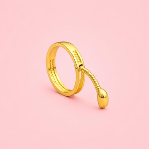 May include: A gold-colored ring with a unique design. The ring features a textured band and a dangling, curved element ending in a rounded shape. The ring is set against a soft pink background.