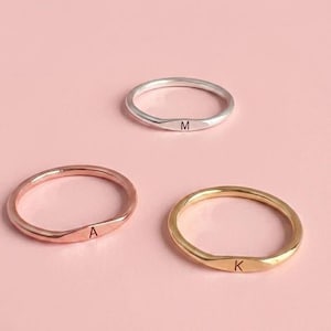 Peut inclure: Trois bagues délicates en argent, or rose et or, chacune gravée d'une seule initiale. Les bagues sont présentées sur un fond rose tendre. Les initiales sont "M", "A" et "K".