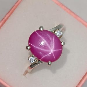 May include: A silver ring featuring a large, oval-shaped, magenta-colored star ruby cabochon as the centerpiece. The gemstone displays a distinct six-rayed star pattern. Two small, round, clear faceted stones are set on each side of the ruby.