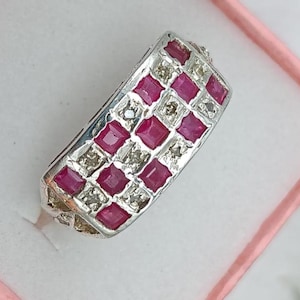 Peut inclure: Une bague argentée présentant un motif en damier composé de pierres précieuses carrées rouge-rose foncé alternant avec de petites pierres transparentes à facettes, disposées en rectangle sur le dessus de l'anneau.