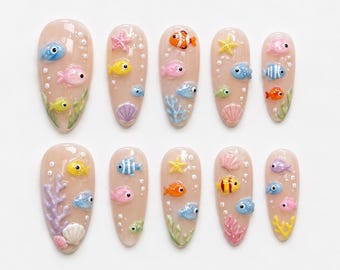 May include: A set of ten almond-shaped press-on nails with a nude base, featuring three-dimensional aquatic decorations including colorful fish, starfish, seashells, and coral accents, along with small white dots representing bubbles throughout.