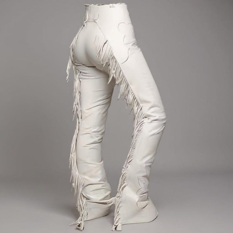 May include: Cream-colored leather chaps with decorative cutouts and long fringe detailing along the sides. The chaps have a high waist and flared leg design, offering a unique Western-inspired aesthetic. The chaps are made of a soft, supple leather.