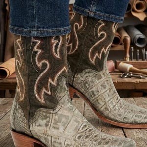 Pode incluir: Um par de botas de cowboy com um padrão de pele de crocodilo texturizado em cinza claro no pé e um cano em verde-oliva escuro. O cano é decorado com bordados intrincados de cor clara em forma de chama. As botas têm um salto de madeira.