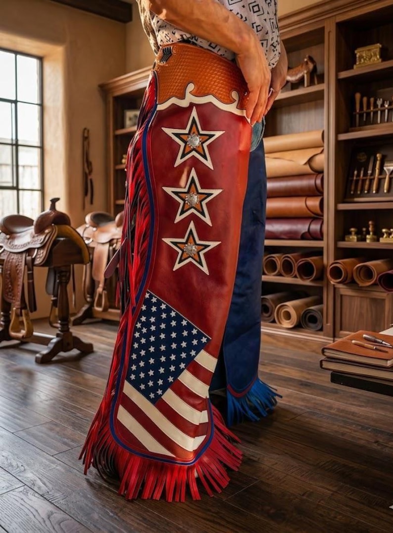May include: Leather chaps featuring a red, white, and blue American flag design on the lower section, three white star appliqu&eacute;s with metallic centers along the side, a textured orange upper panel, and long red fringe trim along the outer leg.