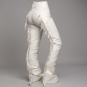 May include: Cream-colored leather chaps with decorative cutouts and long fringe detailing along the sides. The chaps have a high waist and flared bottoms. The overall style is Western-inspired.