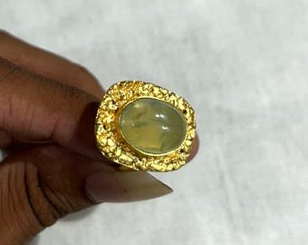 Può includere: Un anello d'oro con una gemma ovale verde chiaro. L'anello ha un'incastonatura in oro strutturata e ornata. L'anello è tenuto su uno sfondo bianco.