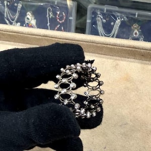 Puede incluir: Un anillo plateado con un diseño calado repetitivo, adornado con pequeñas piedras preciosas transparentes. El anillo es sostenido por una mano enguantada, resaltando sus intrincados detalles. El anillo se presenta sobre una superficie beige, con estuches de joyería en el fondo.