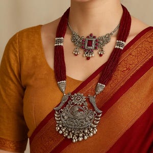 Puede incluir: Una mujer lleva un collar de plata y pendientes a juego. El collar presenta un colgante grande con detalles intrincados y una piedra roja. El collar se combina con un hilo de cuentas rojas. La mujer lleva un top amarillo mostaza y un sari rojo.