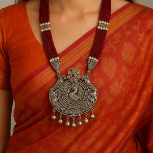 Puede incluir: Un collar con colgante plateado con un diseño de pavo real, hebras de cuentas rojas y pequeños dijes colgantes. El collar se usa sobre un sari naranja.