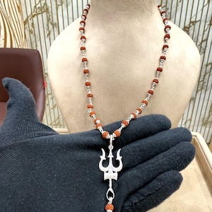 Puede incluir: Un collar de plata con un colgante de tridente y una cadena de cuentas. El colgante tiene un diseño detallado de tres puntas. El collar se exhibe en un maniquí. Las cuentas son de color marrón rojizo.