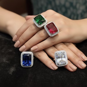 May include: A selection of gemstone rings in green, red, blue, and white. Each ring has a large, rectangular-cut stone with a halo of smaller stones. The rings are displayed on a dark surface. The rings are a mix of colours.