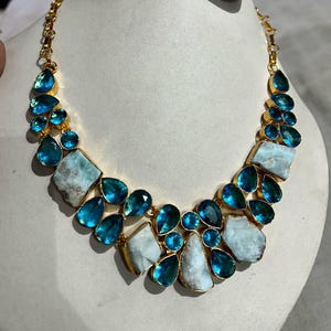 Puede incluir: Un collar dorado con piedras preciosas azules y blancas. El collar presenta piedras azules en forma de lágrima y piedras blancas irregulares. También se ve un par de pendientes a juego con piedras azules cuadradas.