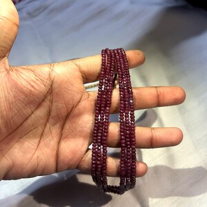 May include: A multi-strand necklace composed of small, round, deep red beads. The necklace is held in a hand, showcasing the color and texture of the gemstones. The beads appear to be strung together, creating a layered effect.