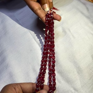 May include: Three strands of deep red, oval-shaped beads are held against a white background. The beads are strung together with white string, and the ends are visible. The beads have a polished, reflective surface.