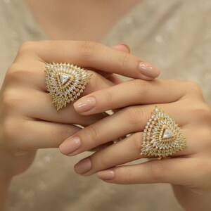 May include: Two gold-toned rings with a triangular design, encrusted with clear, sparkling gemstones. The rings are displayed on a person's hands, showcasing the intricate details of the jewelry. The rings have a central, triangular gemstone.