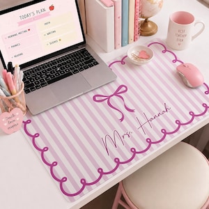 May include: A rectangular desk mat featuring a light pink and white vertical striped pattern with a dark pink scalloped border and a central bow graphic. The mat is personalized with the text Mrs. Hannah in a cursive script font.