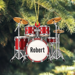 May include: A red drum set ornament featuring a bass drum with the name Robert printed in black text on the white drumhead. The set includes two mounted toms, a floor tom, and two gold-colored cymbals, all suspended by a gold string.