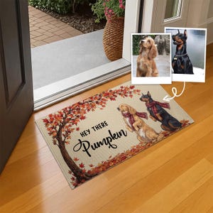 May include: A rectangular doormat featuring a tan background with a large autumn tree with orange and red leaves. Two dogs wearing red plaid scarves are illustrated on the mat. The text "HEY THERE Pumpkin" is printed in black script and block letters.