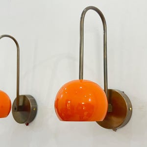 May include: Two wall sconces with orange globe shades and curved metal arms. The sconces have a circular base and are mounted on a white wall. The design is mid-century modern.