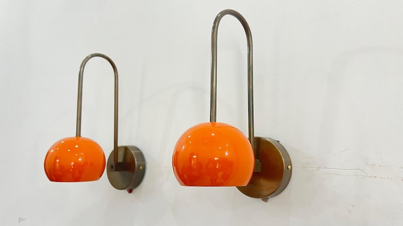 May include: Two wall sconces with orange globe shades and curved metal arms. The sconces have a circular base and are mounted on a white wall. The design is mid-century modern.