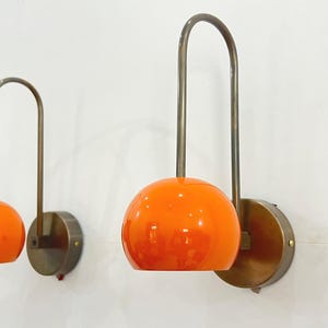 May include: Two wall-mounted lamps with a mid-century modern design. Each lamp features a round, orange glass shade and a curved, metal arm. The lamps are mounted on circular, brass-colored bases. The lamps are against a white wall.