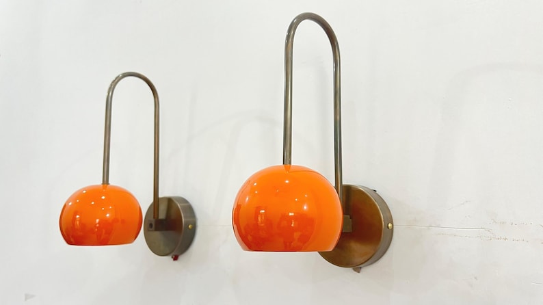 May include: Two wall-mounted lamps with a mid-century modern design. Each lamp features a round, orange glass shade and a curved, metal arm. The lamps are mounted on circular, brass-colored bases. The lamps are against a white wall.