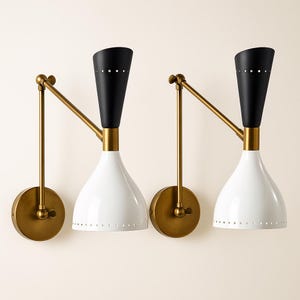 Può includere: Due applique da parete dal design moderno della metà del secolo. Ogni applique presenta un paralume bianco a forma di campana, una parte superiore nera e un braccio e una base color ottone. I paralumi hanno piccoli fori distanziati uniformemente vicino al fondo.