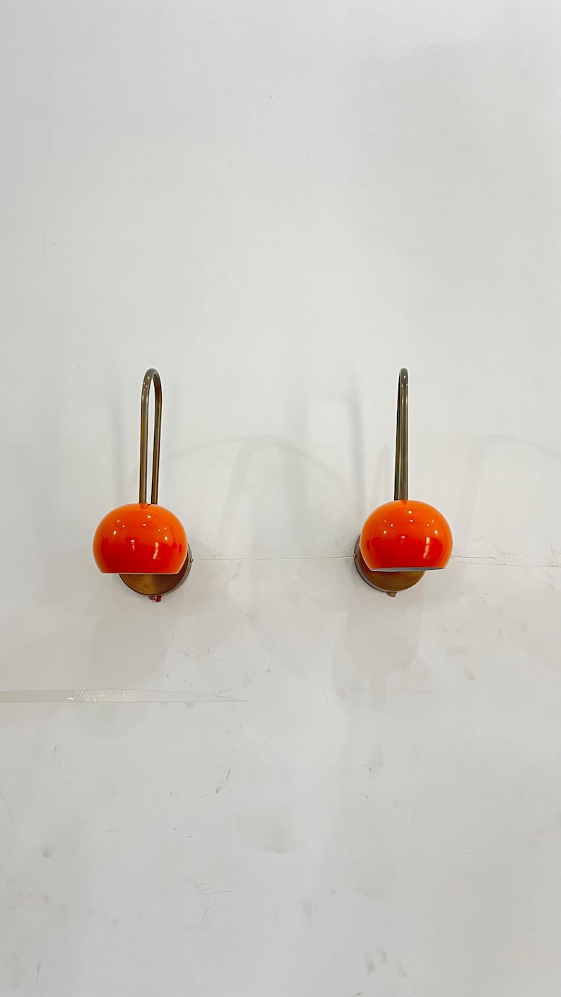 May include: Two wall-mounted lamps with orange, spherical shades. The lamps have a brass-colored, curved arm and a circular base. The lamps are mounted on a white wall, providing a clean and modern aesthetic. The lamps are a decorative lighting fixture.