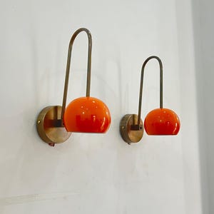 May include: Two wall-mounted sconces with a mid-century modern design. Each sconce features a round, orange glass shade and a curved, brass-toned arm. The shades are mounted on a circular brass base, creating a warm, inviting glow.