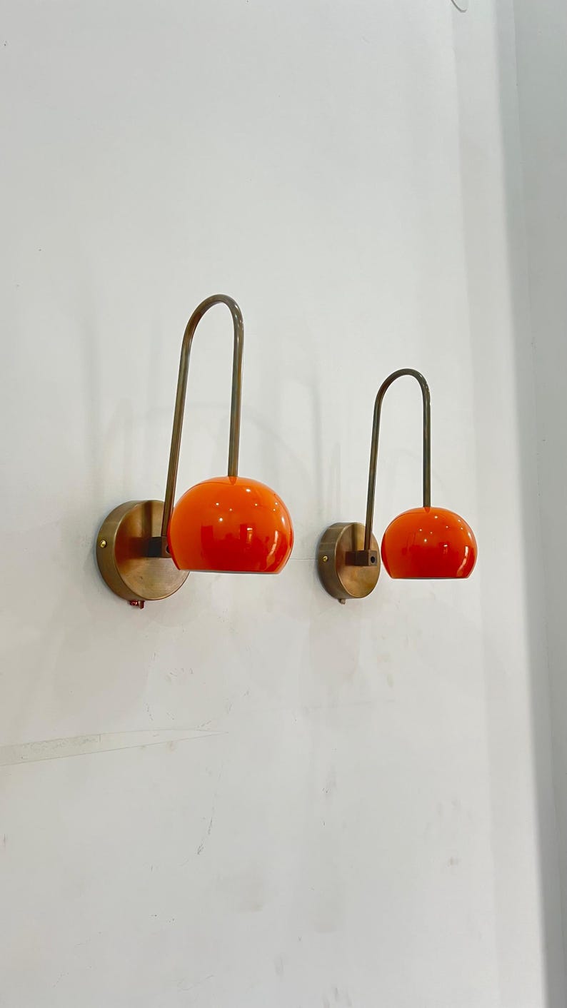 May include: Two wall-mounted sconces with a mid-century modern design. Each sconce features a round, orange glass shade and a curved, brass-toned arm. The shades are mounted on a circular brass base, creating a warm, inviting glow.