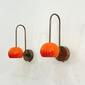 May include: Two mid-century modern wall sconces. Each sconce features a vibrant orange globe shade, a curved brass arm, and a circular brass base. The sconces are mounted on a white wall, offering a pop of color and retro design.