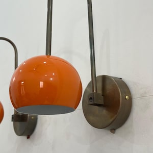 May include: Two wall-mounted sconces with orange globe shades and curved metal arms. The shades are a glossy orange, and the metal fixtures have a bronze finish. The sconces are mounted on round, bronze-colored wall plates.