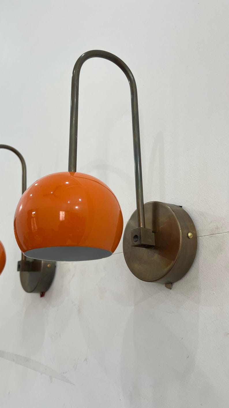 May include: Two wall-mounted sconces with orange globe shades and curved metal arms. The shades are a glossy orange, and the metal fixtures have a bronze finish. The sconces are mounted on round, bronze-colored wall plates.