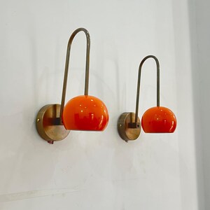 May include: Two wall-mounted sconces with a mid-century modern design. Each sconce features a round, orange glass shade and a curved, brass-toned arm. The shades are mounted on a circular brass base, creating a warm, inviting glow.