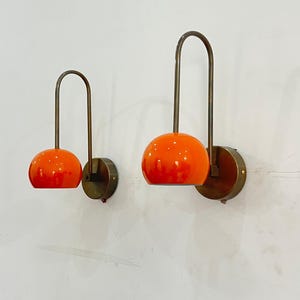 May include: Two wall sconces with orange globe shades and curved brass arms. The sconces are mounted on round brass backplates, creating a mid-century modern aesthetic. The orange shades provide a vibrant pop of color.