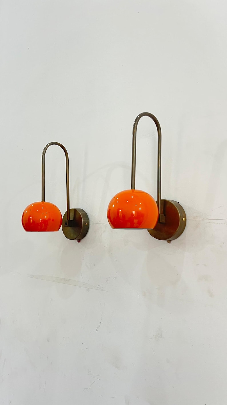 May include: Two wall sconces with orange globe shades and curved brass arms. The sconces are mounted on round brass backplates, creating a mid-century modern aesthetic. The orange shades provide a vibrant pop of color.