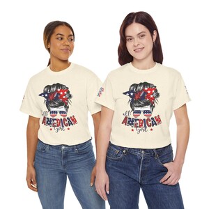 Puede incluir: Una camiseta de manga corta de color crema con un gráfico de un peinado de moño desordenado con un pañuelo de estrellas rojas, blancas y azules, gafas de sol con cristales de bandera estadounidense y el texto "all AMERICAN Girl" en rojo y azul.