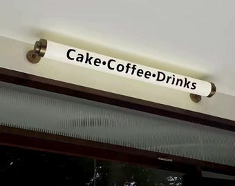 May include: A white, cylindrical light fixture with the words "Cake•Coffee•Drinks" in black lettering. The fixture is mounted on a wall with brass-coloured brackets. The light is positioned above a window.