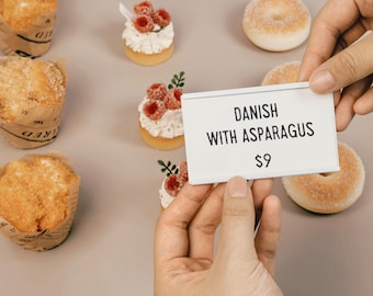 Könnte beinhalten: Eine Auswahl an Backwaren, bestehend aus drei Muffins in Papierförmchen, drei kleinen Törtchen mit Himbeeren und drei einfachen Donuts. Ein in der Hand gehaltenes weißes Schild zeigt DANISH WITH ASPARAGUS $9.