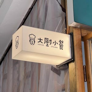 Op de afbeelding: Een rechthoekig, verlicht bord met zwarte tekst en afbeeldingen. Het bord leest "Good Cook Little Meal" in het Engels en Chinese karakters, met een chef-kok icoon. Het bord is gemonteerd op een houten constructie.