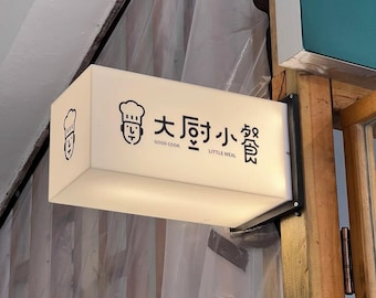 May include: A rectangular, illuminated sign with black text and graphics. The sign reads "Good Cook Little Meal" in English and Chinese characters, with a chef icon. The sign is mounted on a wooden structure.