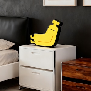 May include: A whimsical yellow and black banana-shaped lamp with a smiling face, resting on a white nightstand. The lamp has a black outline and a yellow body with a curved orange line. The nightstand has two drawers and a silver trim.
